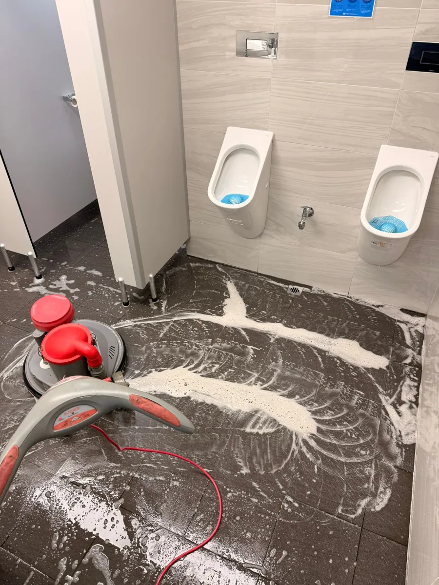 Rotary floor machine scrubbing the tiled floor of an office washroom beside two urinals