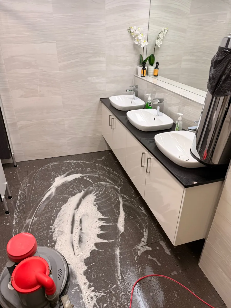 Floor scrubbing machine on cleaning solution along a washroom vanity with three basins