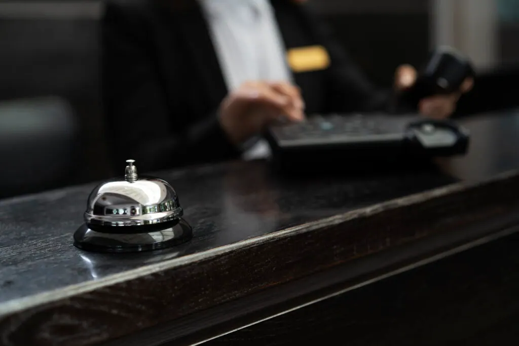A corporate concierge helps an employee at a lobby desk while a hotel bell stands nearby, bridging the workplace and hospitality.