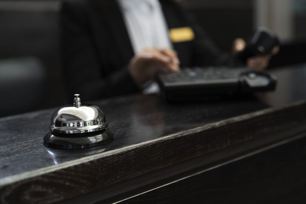 A corporate concierge helps an employee at a lobby desk while a hotel bell stands nearby, bridging the workplace and hospitality.